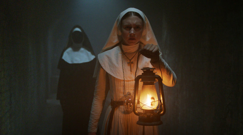 Taissa Farmiga dressed as a nun holding a lantern in a dark hallway while a ghost of anun with no face walks behind her in the movie The Nun