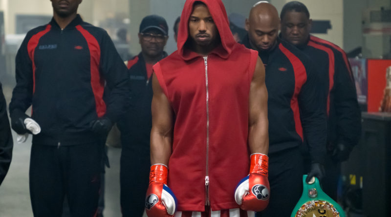 Wood Harris and a team in warmup suits escort Michael B. Jordan as Adonis Creed wearing a red boxing robe in the film Creed II.