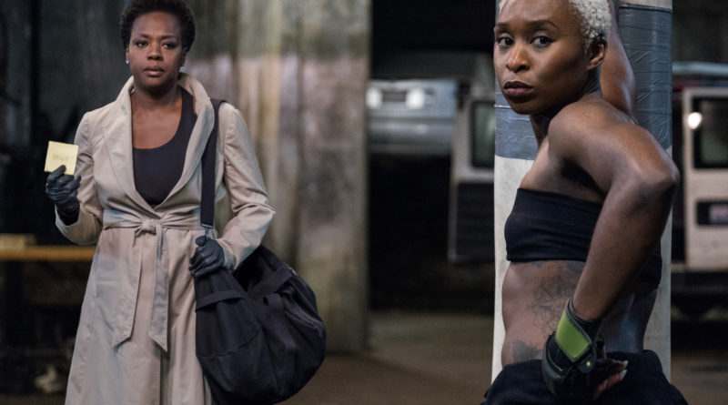 Viola Davis stands in a trenchcoat holding a Post-it note, while Cynthia Erivo stands leaning against a punching bag in workout clothes in the Steve McQueen film Widows