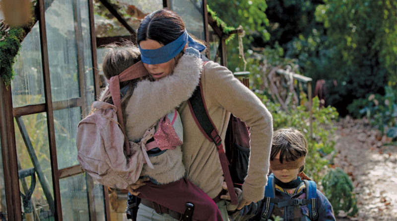 Sandra Bullock blindfolded carrying a girl on her wrapped around her shoulders with a boy walking behind her in the movie Bird Box.